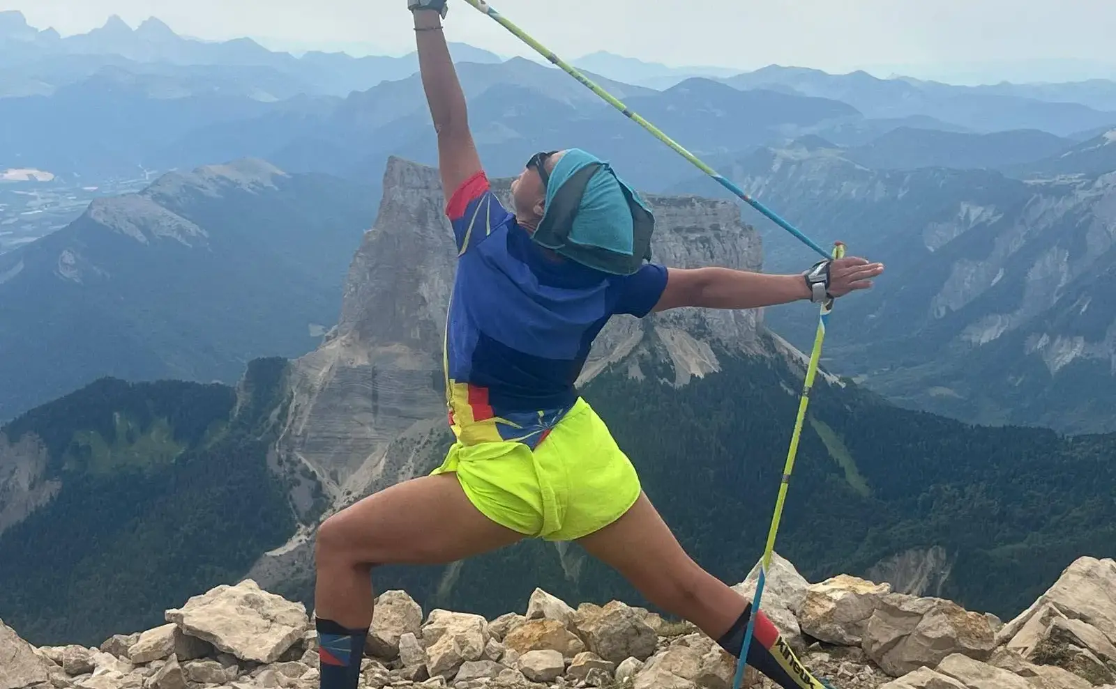 Un pratiquant de Nordic Yoga®, bâtons levés, en posture sur un rocher face au panorama.